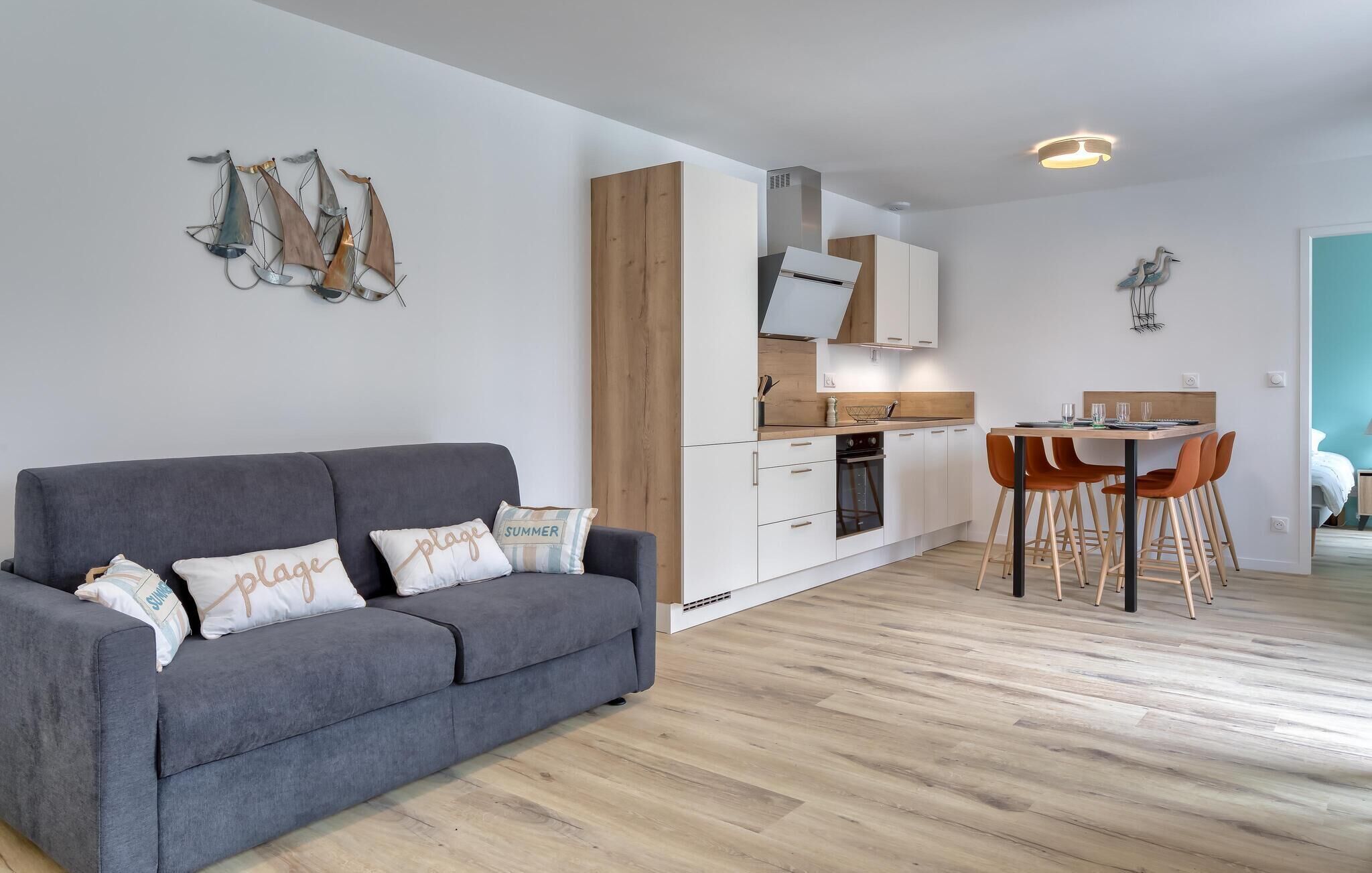 A modern apartment with a grey sofa, wooden flooring, a kitchen with white cabinets, and a dining area with orange chairs.