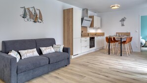 A modern apartment with a grey sofa, wooden flooring, a kitchen with white cabinets, and a dining area with orange chairs. - Gite Gouville sur Mer (Gouville sur Mer)
