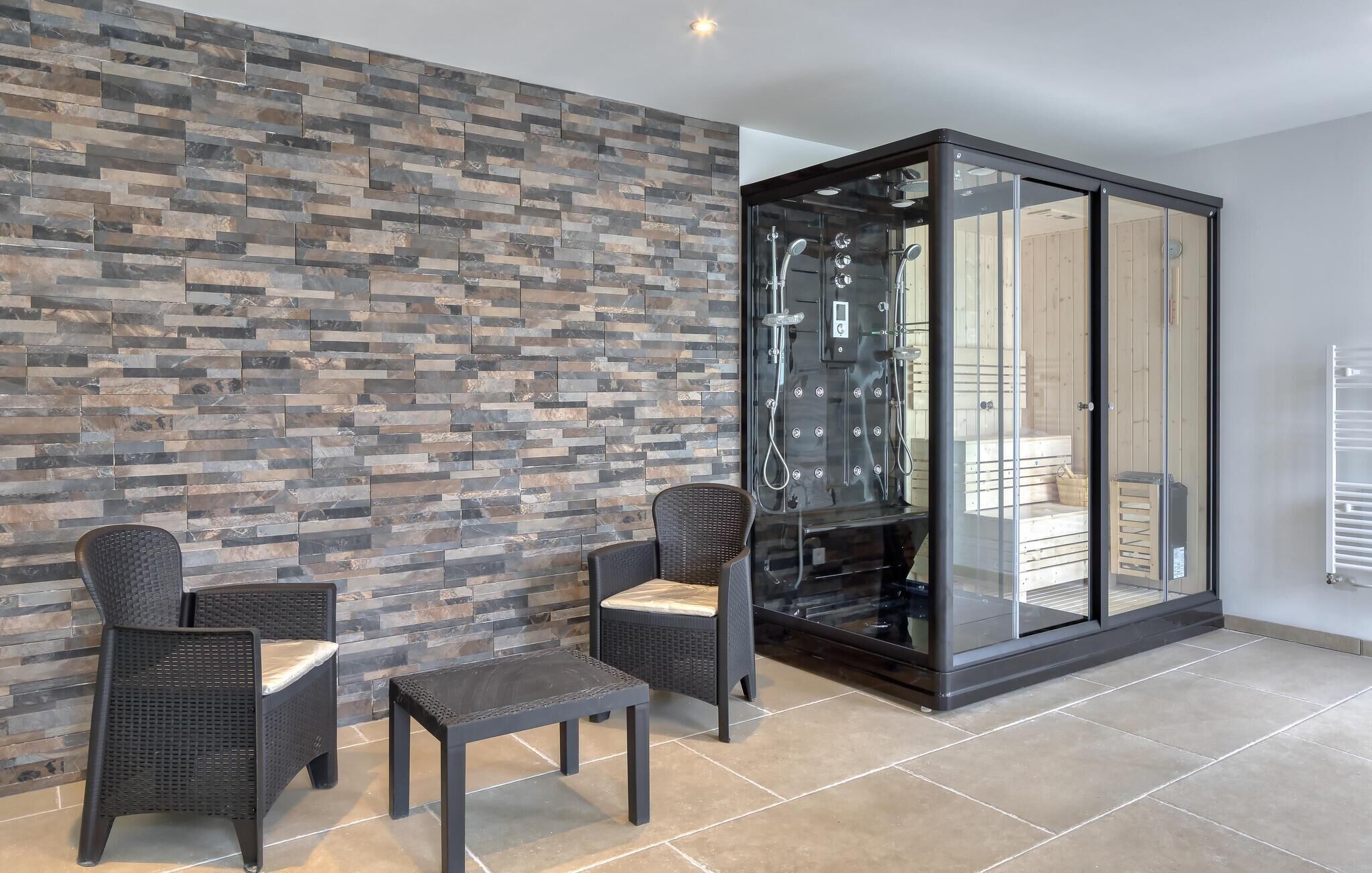 A modern bathroom with a stone wall, two wicker chairs, a small table, and a glass-enclosed shower area.