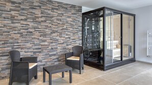 A modern bathroom with a stone wall, two wicker chairs, a small table, and a glass-enclosed shower area. - Gite Gouville sur Mer (Gouville sur Mer)