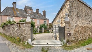 A stone building with a gated entrance, a paved driveway, and a pink house in the background. - Gite Gouville sur Mer (Gouville sur Mer)