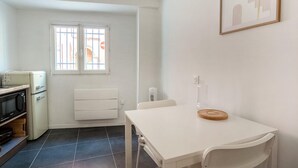 A small, clean kitchen with a white refrigerator, a microwave, and a stove. There is a window with bars, a white radiator, and a white table with a wooden coaster.