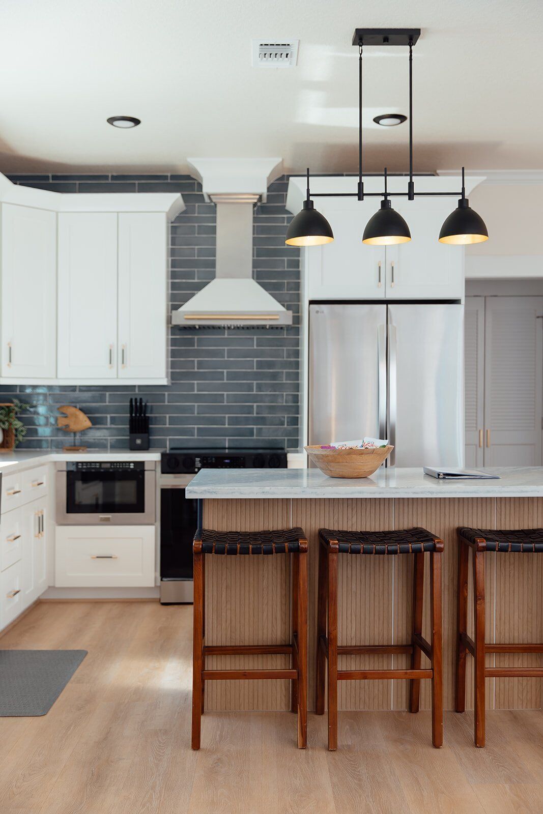 A modern kitchen with white cabinets, a black brick backsplash, stainless steel appliances, and three bar stools with woven seats.