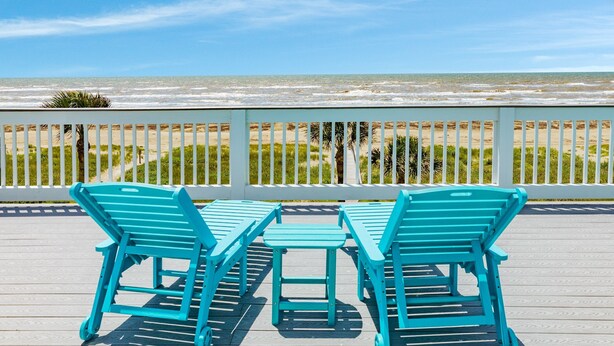 A wooden deck with turquoise chairs and a table, overlooking a beach with palm trees and a fence. - Twin Palms Paradise (Galveston)