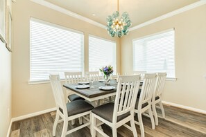 Comedor con mesa de madera, sillas blancas, ventanas amplias con persianas y una lámpara decorativa.