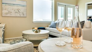 A living room with a round coffee table, a sofa, and a painting of a beach scene. There are three wine glasses and a bottle of wine on the table. - Twin Palms Paradise (Galveston)