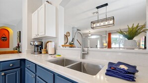 A modern kitchen with white and blue cabinetry, a stainless steel sink, a coffee maker, and a hanging light fixture. - 3 Miles to Disney! Free Heated Saltwater Pool & Spa, Star Wars Game Room, BBQ! (Kissimmee)