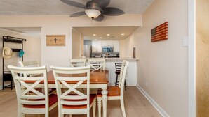 A dining area with a wooden table and white chairs, a ceiling fan, and a kitchen in the background.