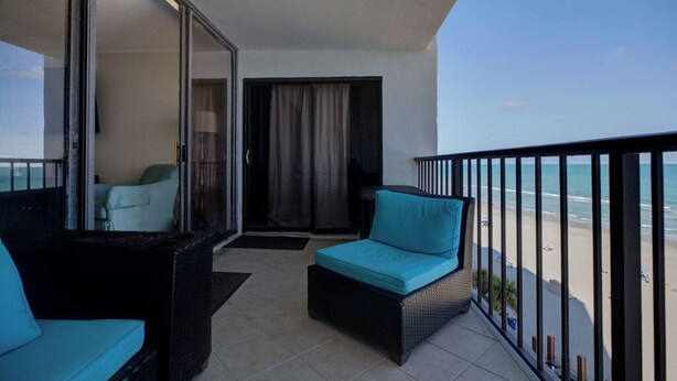 A balcony with a view of the beach, a sofa with a blue cushion, and a chair with a blue cushion.