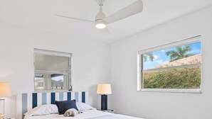 Ein Schlafzimmer mit Bett, Deckenventilator, Nachttisch mit Lampe, Fenster mit Blick auf Grünflächen und Spiegel an der Wand.