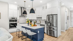 A modern kitchen with white cabinets, a stainless steel refrigerator, and a blue island with bar stools. - Luxury Oceanside Escape – Balcony, Patio & Steps to Indialantic Beach! (Indialantic)