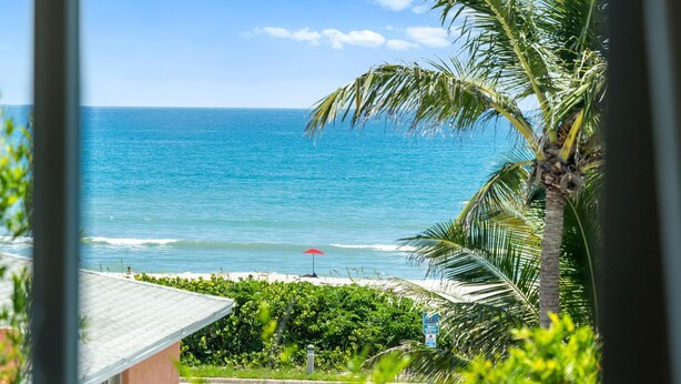 A view of a beach with a clear blue ocean, a red umbrella, and palm trees. - Luxury Oceanside Escape – Balcony, Patio & Steps to Indialantic Beach! (Indialantic)