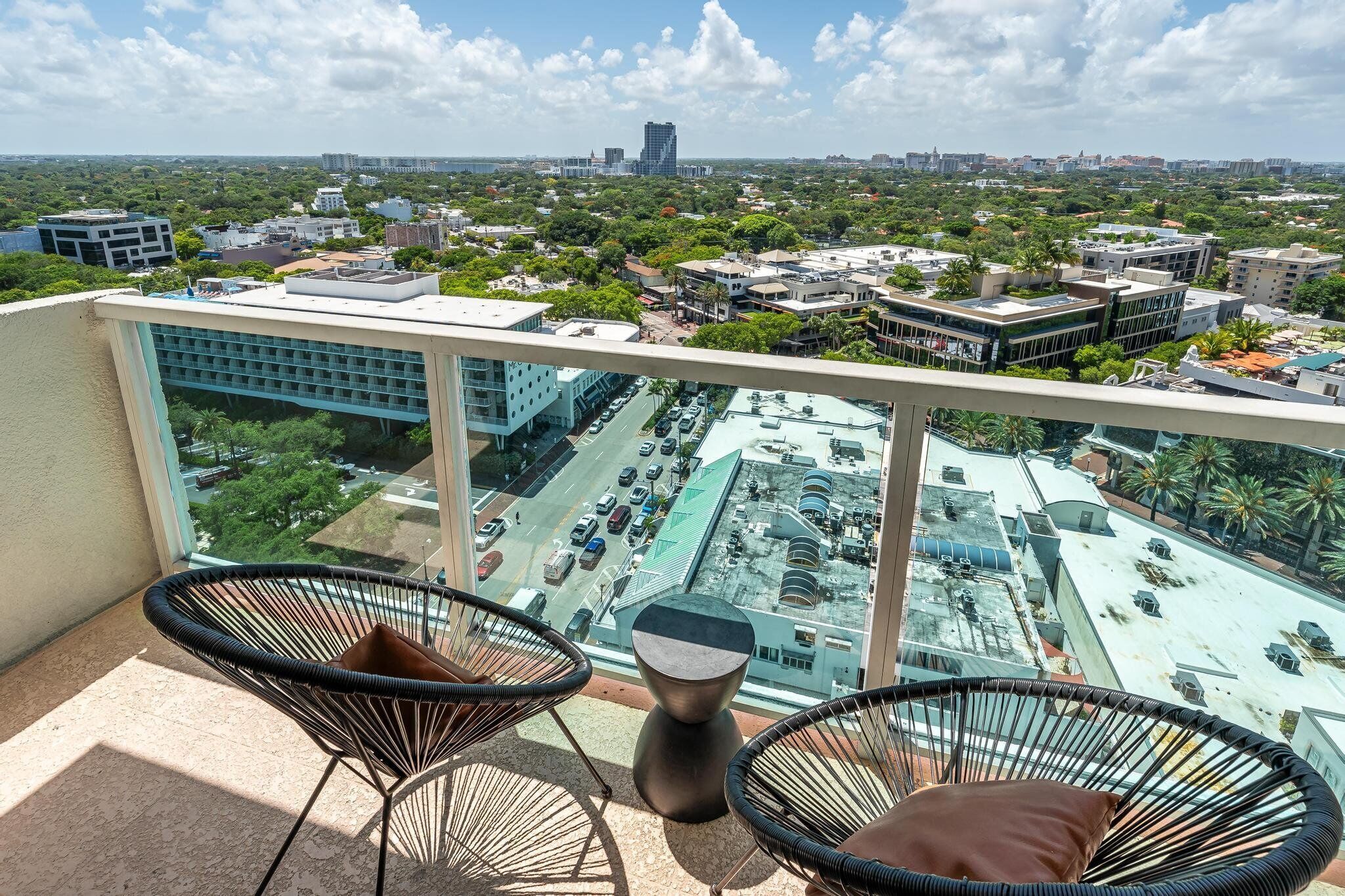 A balcony with two wicker chairs and a small table, overlooking a parking lot and buildings with a clear sky.