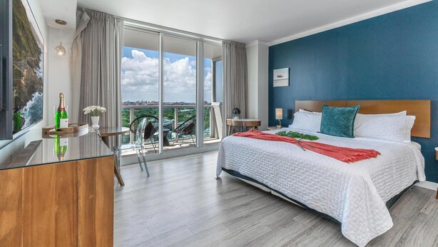 A modern hotel room with a large bed, a desk, and a balcony with a city view.