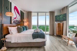 A modern hotel room with a large bed, a TV, and a balcony with a city view. - Modern 3BR/3.5BA Grove Condo | Views, Balconies, Full Kitchen + 3 Parking (Miami)