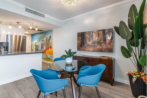 A modern kitchen with stainless steel appliances, a glass dining table with blue chairs, and a large painting of a tropical scene. - Modern 3BR/3.5BA Grove Condo | Views, Balconies, Full Kitchen + 3 Parking (Miami)