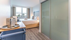 A hotel room with a bed, a small kitchenette with a kettle, a sliding glass door, and a view of palm trees.
