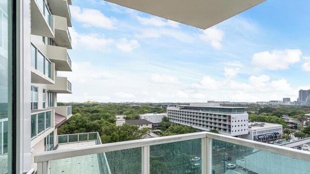 A high-rise building with multiple balconies, large windows, and a view of a cityscape with greenery and other buildings.