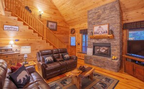 A log cabin interior with a fireplace, leather sofa, wooden staircase, and a television.