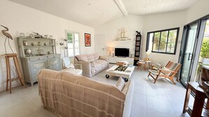 A cozy living room with a sofa, a coffee table, and a TV. There's a bookshelf and a window with a view.