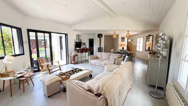 A spacious living room with a large sofa, a coffee table, and a dining area with chairs. There are large windows allowing natural light and a view of the outdoors.