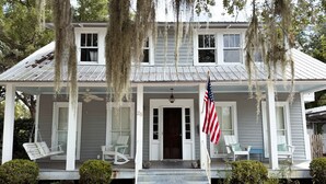 A two-story house with a porch, a flag, and a swing chair. - Historic Coastal Retreat: 3‑BR Cape Cod Cottage w/ Courtyard, Porch & Bay Access (Apalachicola)