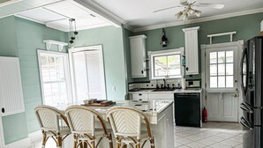 A kitchen with a central island, white cabinetry, and a dining area with wicker chairs. - Historic Coastal Retreat: 3‑BR Cape Cod Cottage w/ Courtyard, Porch & Bay Access (Apalachicola)