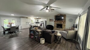 Een ruime woonkamer met een open haard, een eethoek met een tafel en stoelen, en een keuken met witte kasten en een accentmuur van steen.