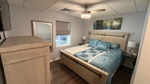 Een slaapkamer met een bed, nachtkastje, dressoir, raam met jaloezieën, plafondventilator en een schilderij aan de muur.