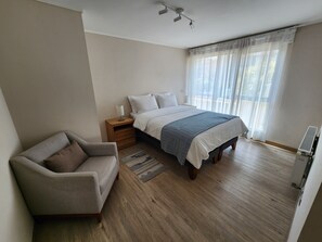 Une chambre à coucher comprenant un lit, une table de chevet, une chaise, une fenêtre avec des rideaux légers et un ventilateur de plafond.