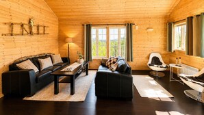 Interior de una cabaña de madera con un sofá y un sillón de cuero negro, una silla blanca, una mesita y una ventana con cortinas.
