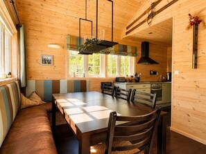 Un intérieur en bois comprenant une table à manger, des chaises, un banc, un coin cuisine et une fenêtre donnant sur un paysage verdoyant.