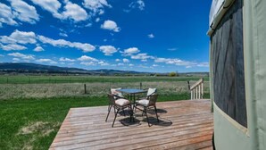 A wooden deck with a table and chairs, overlooking a grassy field and distant hills under a partly cloudy sky. - Enchanting yurt in secluded mountain view setting (Swan Valley)