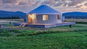 Une yourte dotée d’une porte et de fenêtres en bois, située dans un champ verdoyant avec des montagnes en arrière-plan.