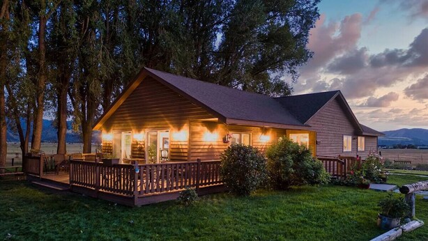 Une cabane en bois avec un porche, entourée d’arbres et bordée d’une pelouse.
