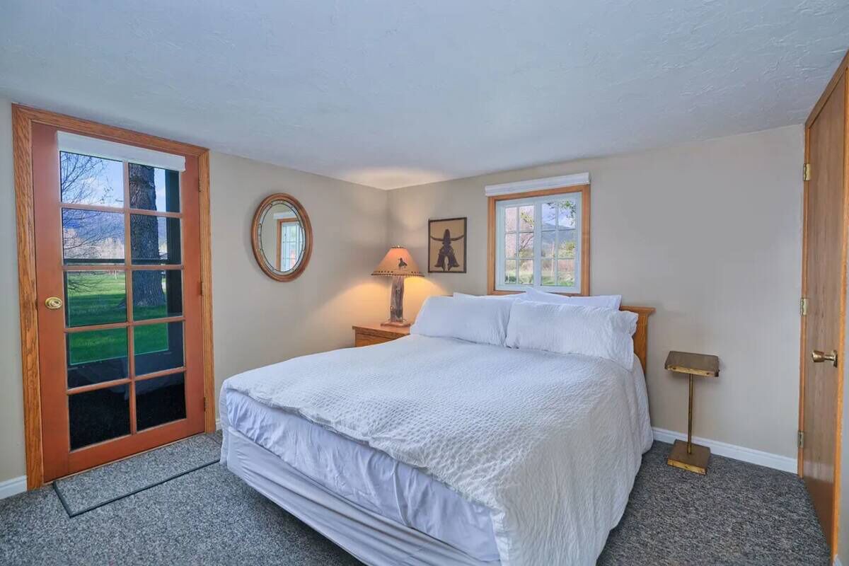 A bedroom with a large bed, a wooden door, a window, a bedside table with a lamp, and a framed picture on the wall.