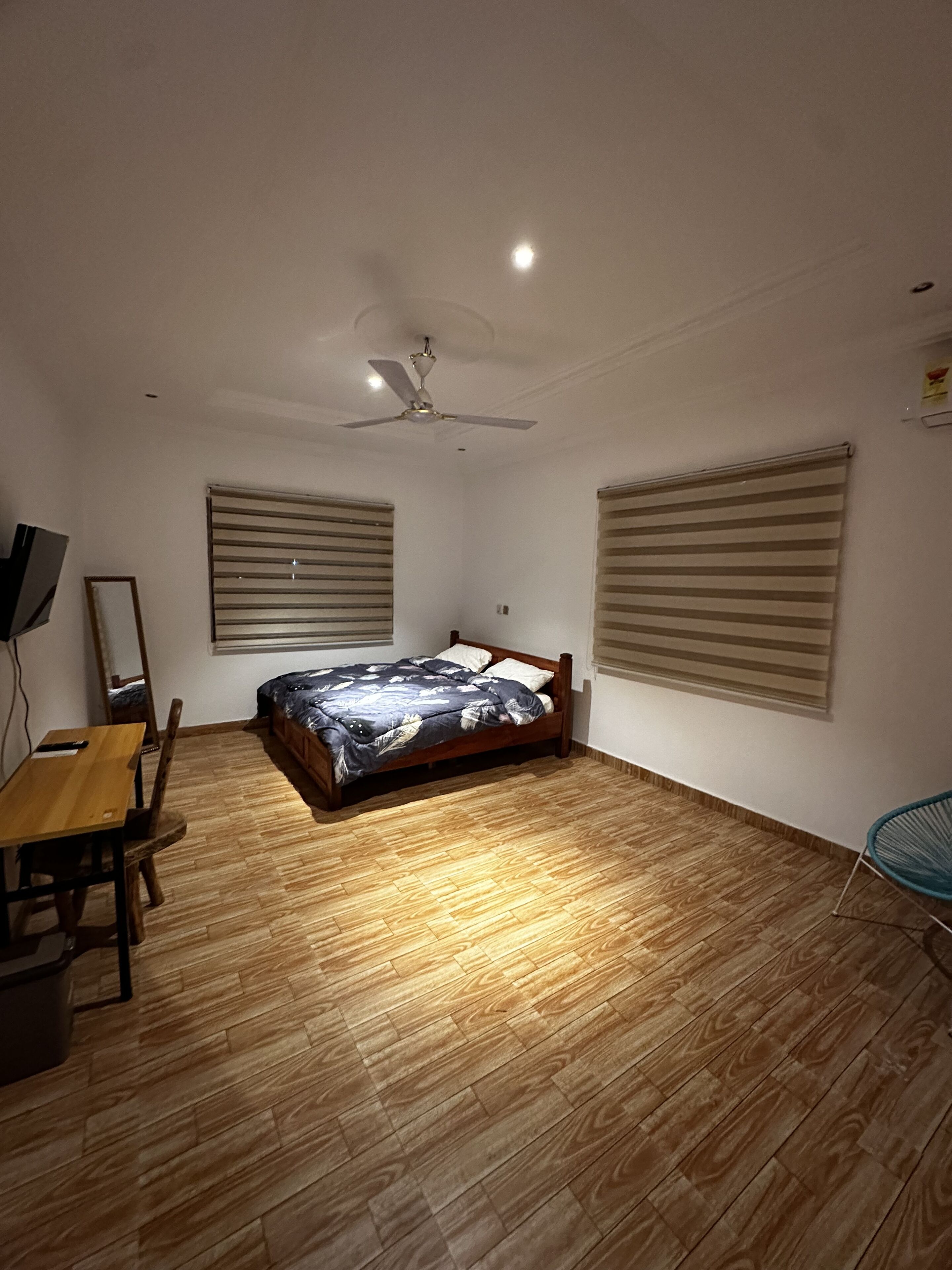 Een slaapkamer met een houten vloer, een bed met een beddeken met een patroon, een plafondventilator en twee ramen met jaloezieën.