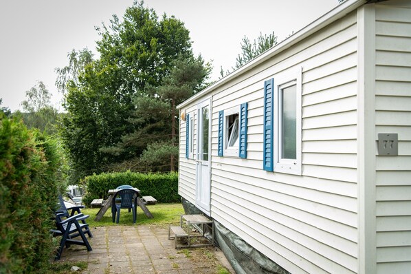 A white mobile home with blue shutters, a patio with chairs, and a small table. - Mobile Home With Heated Pool (Manhay)