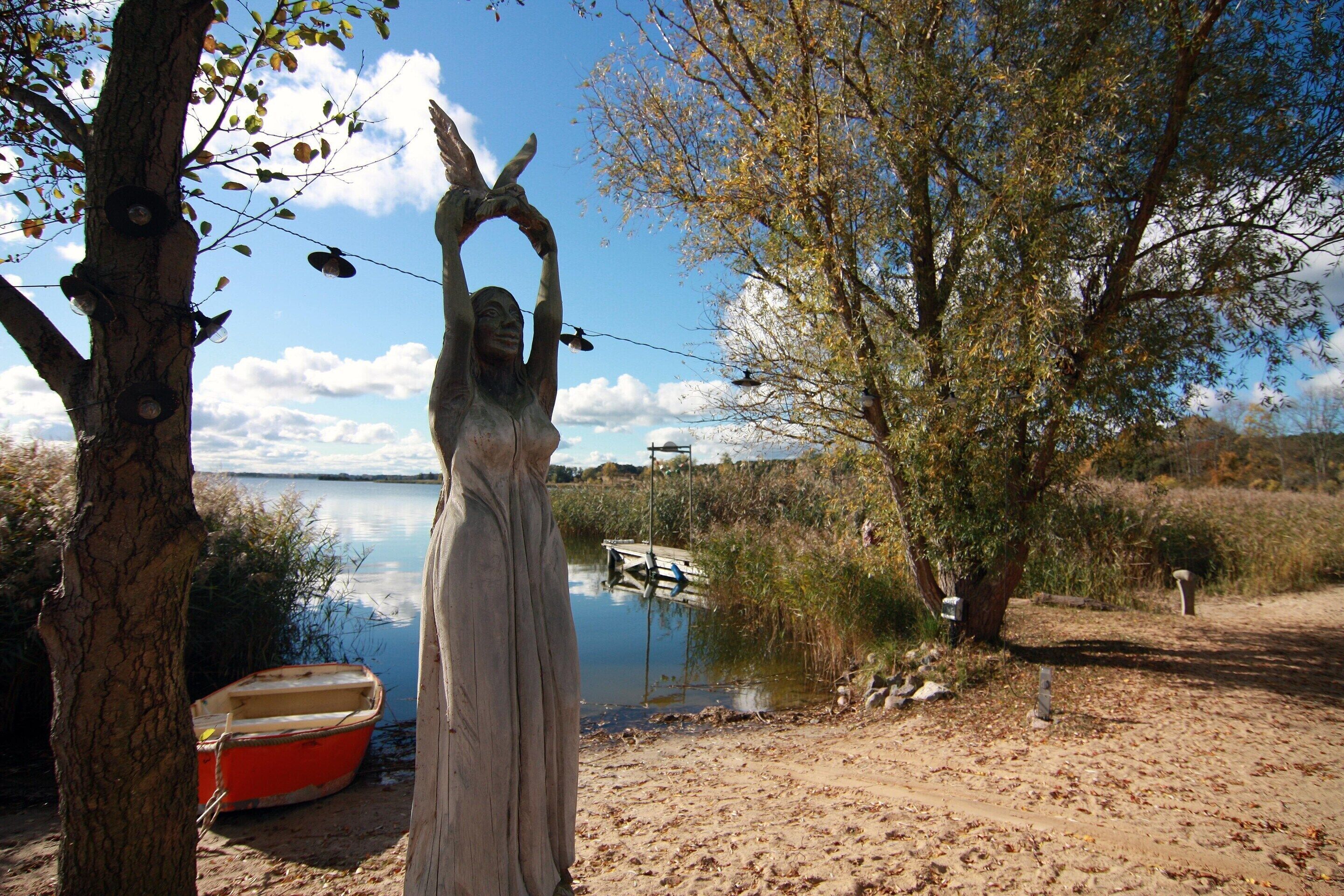 Una estatua de una persona con las manos levantadas cerca de un cuerpo de agua, un bote y árboles en un entorno natural.