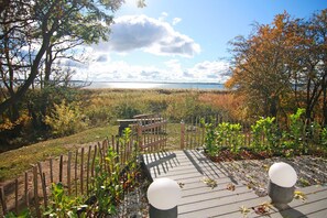 Eine Holzterrasse mit einer Bank, umgeben von einem Zaun und Bepflanzung, mit Blick auf ein Gewässer.
