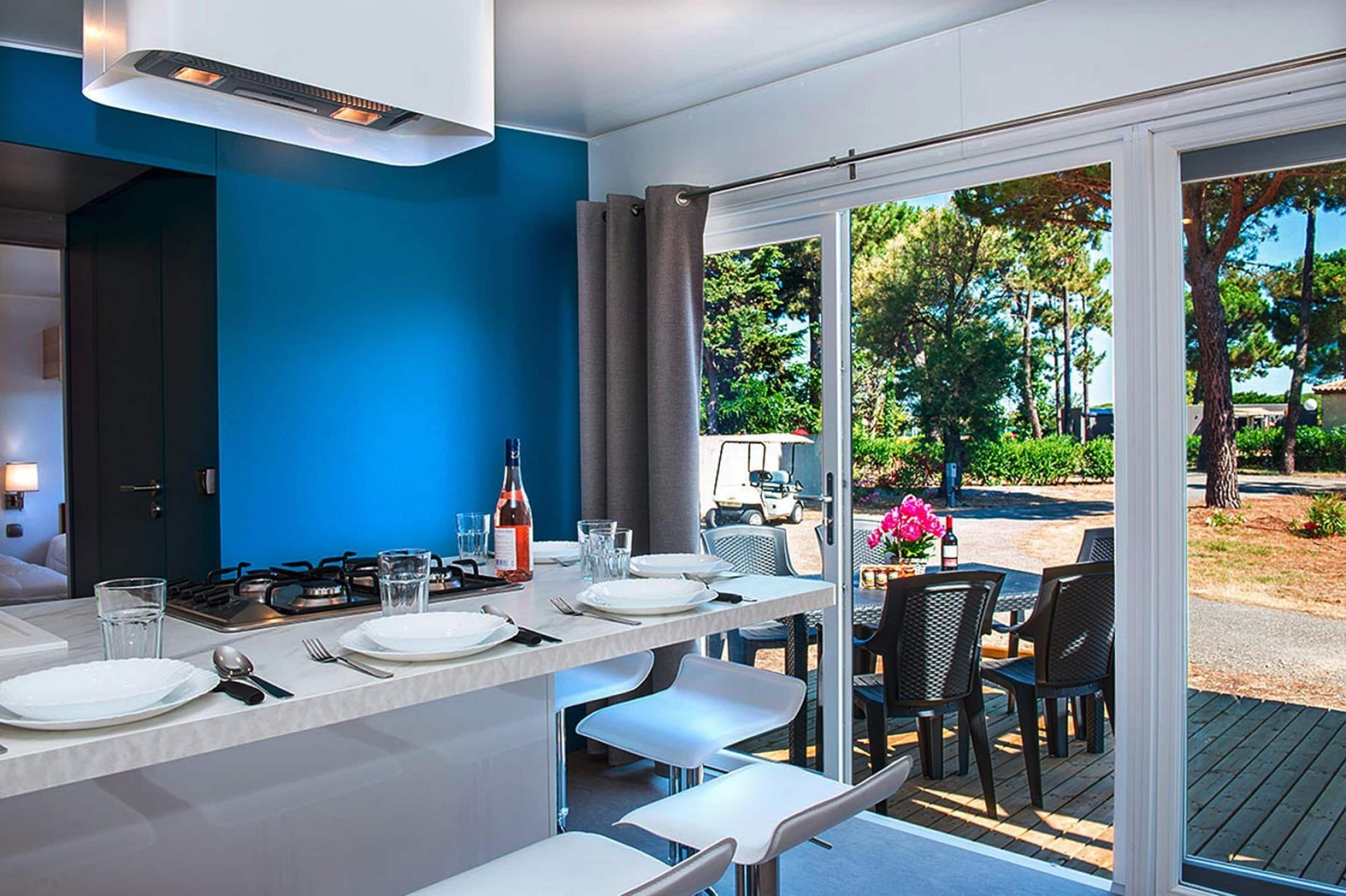 A modern kitchen with a blue accent wall, a dining area with white chairs, and a view of a garden through large sliding doors.