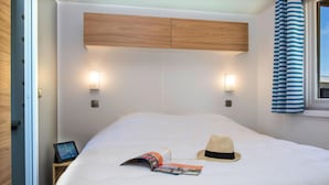 Een netjes opgemaakt bed met een tijdschrift en een stroohoed erop, een nachtkastje met een tablet, en twee wandlampen.
