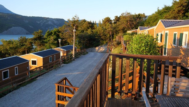 A wooden deck leading to a gravel path with cabins and a view of a lake and mountains. - Apartment on Lake Serre-poncon With Pool (Chorges)