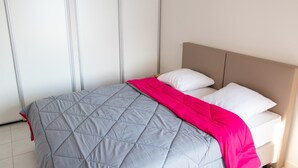 A neatly made bed with a grey quilt and a pink throw blanket, white pillows, and a wooden headboard in a modern room with white walls and a tiled floor. - Apartment on Lake Serre-poncon With Pool (Chorges)