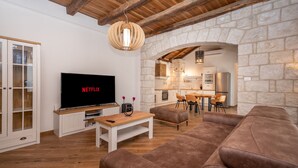 A cozy living room with a television displaying 'NETFLIX', a stone wall, wooden furniture, and a kitchen area in the background.