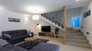 A modern living room with a grey sofa, a coffee table, a TV on a wooden stand, a staircase with wooden steps, and a painting on the wall.