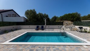 Ein rechteckiger Swimmingpool mit Fliesen umgeben, zwei Liegestühle, ein Sonnenschirm und ein Grill.