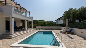 A rectangular swimming pool with a metal ladder, surrounded by a tiled patio. There are lounge chairs and a covered seating area with a table and chairs. The pool is adjacent to a building with a pergola and a view of green hills in the background.