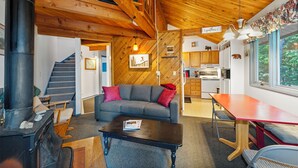 A cozy cabin interior with a wooden staircase, a sofa with red cushions, a dining table, and a kitchen area. - New! Spruce Nest Cabin - Alpine Skiing | Community Pool and Hot Tub (Whitefish)
