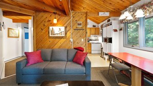 A cozy living room with a wooden ceiling, a gray sofa with red cushions, a dining table, and a kitchen area in the background. - New! Spruce Nest Cabin - Alpine Skiing | Community Pool and Hot Tub (Whitefish)